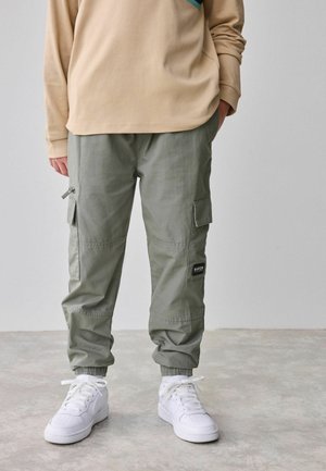 Pantaloni cargo dintr-un material verde măsliniu deschis, cu manșete elastice, buzunare laterale și o etichetă cu numele brandului. Asortați cu adidași albi.