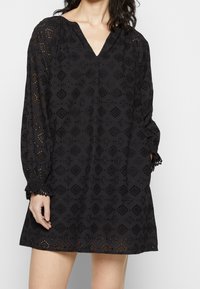 Donna che indossa un vestito nero a maniche lunghe, lungo fino al ginocchio, con motivi in pizzo occhiello e un scollo a V, mentre posa con una mano in tasca.