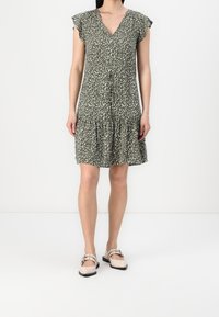 Grünes Blumenkleid mit V-Ausschnitt, kurzen rüschierten Ärmeln und einem gestuften Saum. Kombiniert mit hellen flachen Schuhen, die eine dekorative Schnalle haben.