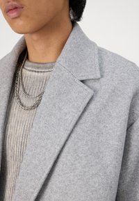 Cappotto grigio in misto lana con colletto a punta, abbinato a un maglione grigio chiaro a coste e collane in argento a strati.