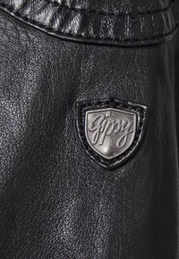 Black faux leather material featuring a stitched patch with a silver logo embossed in a shield shape. Smooth texture with subtle shine.