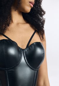 Corsetto in pelle nera con coppe imbottite, caratterizzato da sottili spalline nere e una texture liscia. Ha un design strutturato e dettagli delle cuciture.