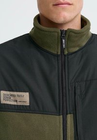 Giacca zip-up verde e nera con colletto in pile, caratterizzata da un patch etichettato 'Dedicato alla Perfezione del Denim' e una tasca frontale.