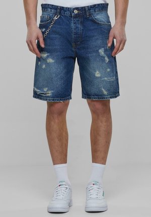 Donkerblauwe denim shorts met versleten plekken, vijf zakken, riemlussen en een metalen kettingaccent, gecombineerd met witte sneakers en sokken.