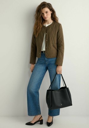 Kobieta ubrana w oliwkową pikowaną kurtkę i niebieskie dżinsy trzyma czarną skórzaną torbę typu tote, stojąc na tle jednolitego jasnego tła.