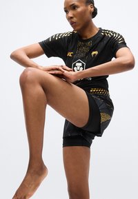 Črni športni dres z zlatimi poudarki in teksturiranim vzorcem. Hlače so črne z geometričnim vzorcem. Oseba je v atletski pozi.