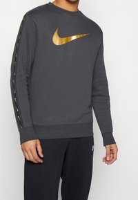 Tmavě šedá mikina Nike se zlatým logem na hrudi a zlatými akcenty na rukávech. Měkká, sportovní látka.