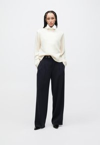 Cream, ribbed knit sweater with high neckline, paired with high-waisted, dark blue wide-leg trousers and black pointed-toe shoes.