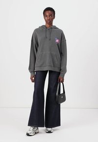 Haut à capuche gris surdimensionné avec une poche avant et un patch logo coloré, associé à un jean en denim foncé évasé et des baskets noires, tenant un petit sac.
