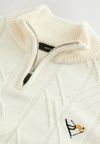 Adīts, krēmkrāsas pus-zip dšers ar ribinātu apkakli. Iezīmēts ar rakstainu tekstūru un mazu izšūtu slēpotāja detaļu uz krūtīm.