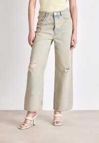 Light blue wide-leg jeans with distressed rips, high waist, and raw hem; paired with cream strappy heels. Smooth denim texture.