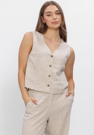 Vrouw draagt een beige mouwloos vest met knopen en bijpassende broek, staat met handen in de zakken tegen een effen witte achtergrond.