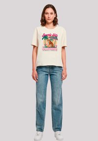Weißes Baumwoll-T-Shirt mit einem bunten Grafikdesign, das einen Tiger und Palmen zeigt, kombiniert mit hellblauen Jeans und weißen Sneakers.