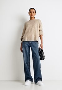 Béžový, prehnaný pletený sveter s rolovanými rukávmi, kombinovaný so svetlomodrými širokými džínsami. Drží malú čiernu tašku s bielymi bodkami.