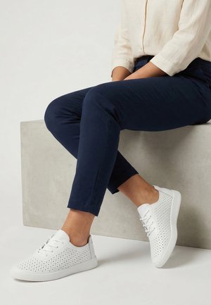 Osoba siedząca na betonowym bloku, ubrana w granatowe spodnie i białe perforowane sneakersy ze sznurówkami na jasnym tle.