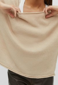 Beżowy sweter dzianinowy o luźnym kroju, z szerokim dekoltem i ściągaczem. Osoba lekko naciąga materiał.