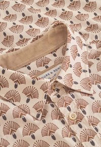 Camicia di cotone beige con un motivo a ventaglio marrone, con piccoli accenti neri e colletto a bottoni. Leggera texture, design casual.