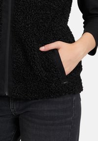 Czarna kamizelka z polaru o fakturowej tkaninie, z zamkiem błyskawicznym i kieszeniami po bokach. Połączona z czarnymi dżinsami, tworzy monochromatyczny wygląd.