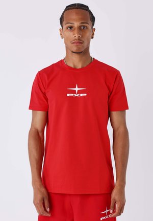 Jeune homme aux cheveux tressés portant un T-shirt rouge avec le logo PXP et un pantalon rouge assorti, debout devant un fond blanc uni.