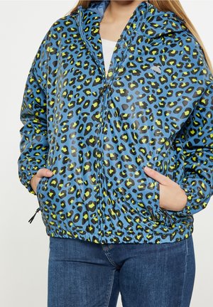 Veste bleue zippée avec imprimé léopard jaune et noir portée par une personne les mains dans les poches, associée à un jean bleu foncé.