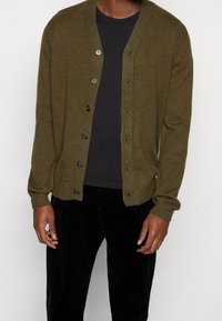 Olivgrön kofta, stickad textur, knäppning i fram med fyra bruna knappar, ribbad krage, bärs över en svart t-shirt, i kombination med svarta byxor.