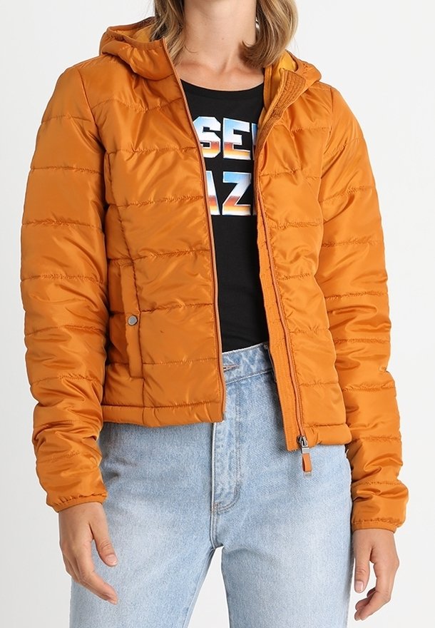 Orange vadderad jacka med huva, med dragkedja framtill, sidofickor och quiltad textur. Bärs över en svart skjorta och ljusa jeans.