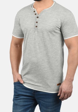 Mężczyzna w jasnoszarej koszulce henley z krótkim rękawem z czterema guzikami, białym wykończeniem i niebieskich dżinsach, z ręką w kieszeni na jednolitym tle.