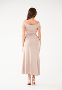 Abito lungo bodycon in beige chiaro, con scollatura off-the-shoulder e dettagli arricciati. Tessuto morbido e stretchy, abbinato a tacchi a strappy.