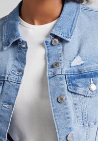 Person som bär en ljusblå sliten denimjacka med metalldetaljer över en vit ribbad skjorta.