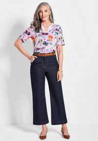 Kortslachtige, multicolor bloementop met een witte kraag, gecombineerd met hoge taille, donkere spijkerbroek met brede pijpen en bruine hakken.