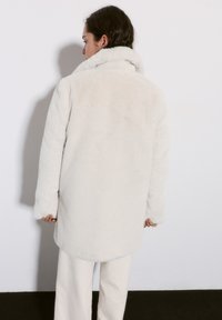 Fehér műszőrme kabát magas gallérral, hosszú ujjakkal és térd közepéig érő egyenes szabással. Texturált felület és varrás nélküli kialakítás.