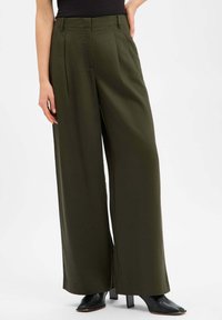 Wide-leg trousers in dark olive green fabric, featuring pleats, clean lines, and a smooth texture. Paired with pointed black heels.