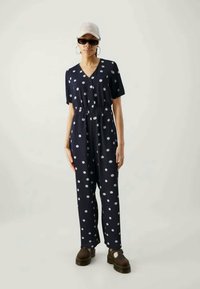 Μπλέ τζάκετ με πουά σχέδιο, κοντά μανίκια, V-λαιμόκοψη και ελαστική μέση. Κατασκευασμένο από ελαφρύ ύφασμα, συνδυασμένο με καφέ πλατφόρμες.