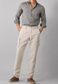 Man in een grijs lange mouwen shirt met opgerolde mouwen, beige cargo broek en bruine casual schoenen, staand met handen in de zakken.