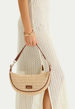 Mano sosteniendo un bolso tejido beige con ribete y correa marrones, combinado con un vestido de crochet crema y una pulsera de oro.