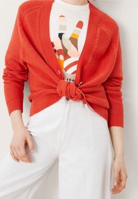 Röd ribbad cardigan knuten i midjan över en vit t-shirt med grafik. Ljust färgade vida jeans med en slät textur.