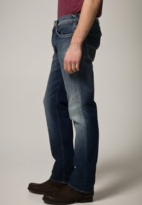Jeans de denim con corte tapered; presentan un lavado degradado de azul oscuro a azul claro; diseño de cinco bolsillos; llevados con zapatos marrones.