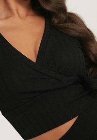 Nahaufnahme einer Frau in einem schwarzen, gerippten Wickeloberteil mit V-Ausschnitt und langen, dunklen, welligen Haaren vor einem neutralen Hintergrund.