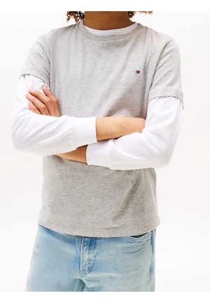 Person med krøllet hår iført en grå kortærmet skjorte over en langærmet hvid skjorte og lyseblå jeans, med armene krydset.