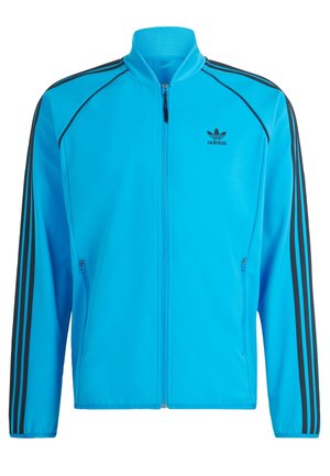 Giacca sportiva blu con strisce nere. Presenta una chiusura a zip frontale, colletto rialzato e due tasche laterali con zip. Realizzata in materiale morbido e leggero.