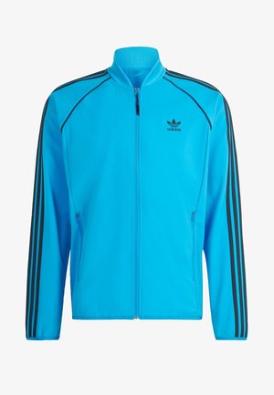 Giacca sportiva blu con strisce nere. Presenta una chiusura a zip frontale, colletto rialzato e due tasche laterali con zip. Realizzata in materiale morbido e leggero.