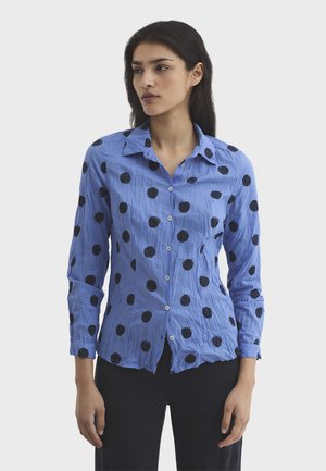 Kobieta w pogniecionej, niebieskiej koszuli na guziki z dużymi czarnymi grochami i czarnych spodniach, stojąca na tle jednolitego jasnego tła.