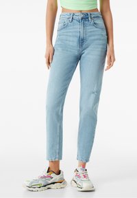 Högmidjade ljusblåjeans med slitna detaljer, i kombination med flerfärgade sportskor som har accenter i rosa, orange och grönt.