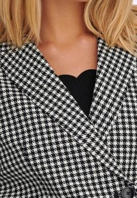 Blazer s černobílým houndstooth vzorem, který má klopový límec, texturovanou látku a zapínání na jeden knoflík.