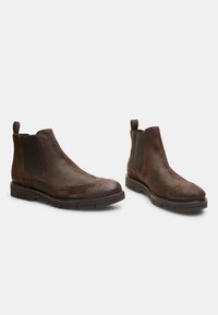 Bottines en cuir marron avec des panneaux élastiques sur les côtés, des détails brogue et une semelle en caoutchouc robuste ; elles disposent d'une sangle à l'arrière pour un enfilage facile.