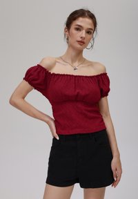 Rød av-shoulder topp med teksturert mønster og puffede korte ermer, kombinert med svarte denimshorts. Sølvhalskjede som aksent.