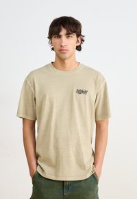 Beige cotton t-shirt with a classic crew neck and short sleeves. Features a small printed design on the left chest and a relaxed fit.