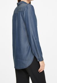Denim shirt in dark blue, featuring a curved hem, long sleeves with buttons, and a subtle seam detail on the back.