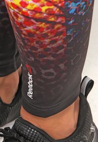 Leggings Reebok con un motivo esagonale colorato, principalmente arancione, rosso e blu, con una base in tessuto nero e dettagli del marchio in bianco.