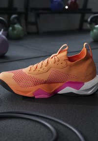 Chaussure d'entraînement athlétique orange et rose posée sur le sol de la salle de sport avec des kettlebells et une corde en arrière-plan flou.